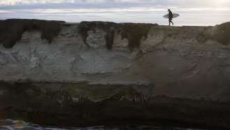 A surfer walks a rock ledge near the ocean.