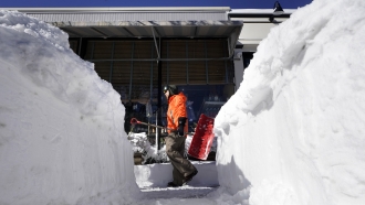A man carries a snow shovel past a snow bank on a sidewalk