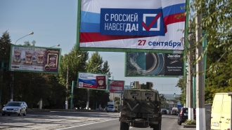 A military vehicle drives along a street with a billboard that reads: 