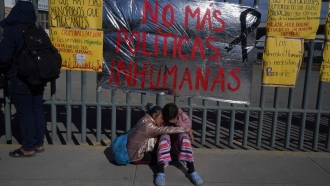 A pair of Venezuelan sisters comfort each other sitting on a sidewalk outside an immigration detention center.