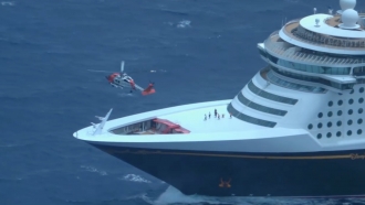 A helicopter sits in the air above a cruise ship.