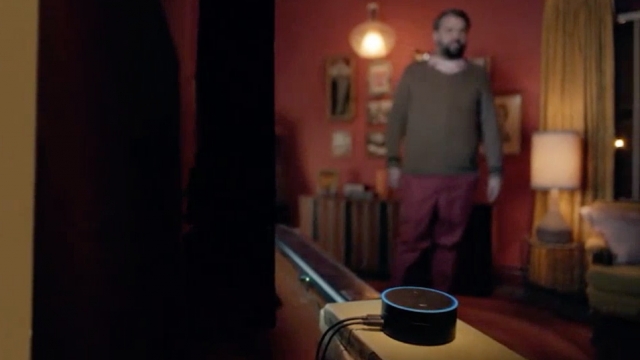 Amazon's Echo Dot lights up in the foreground while a man nearby speaks to the device.