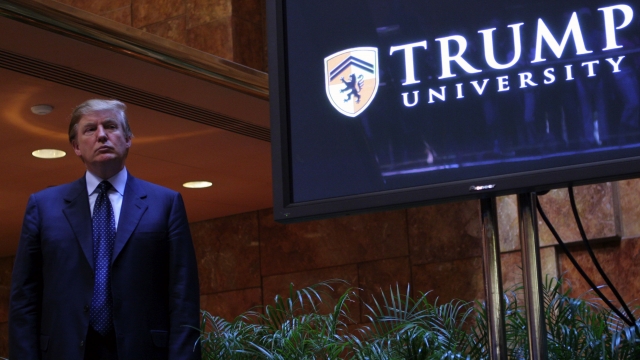 Donald Trump stands next to a TV displaying the Trump University logo.
