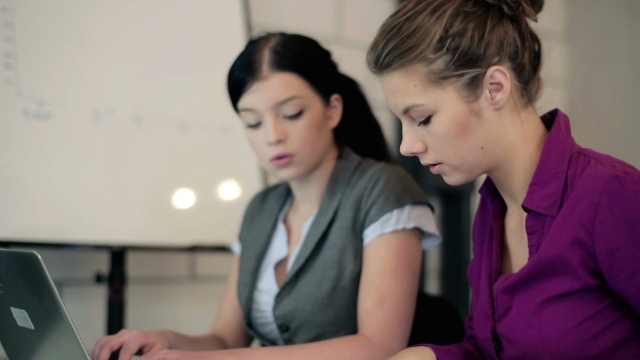 Two young women work on a laptop in an office setting