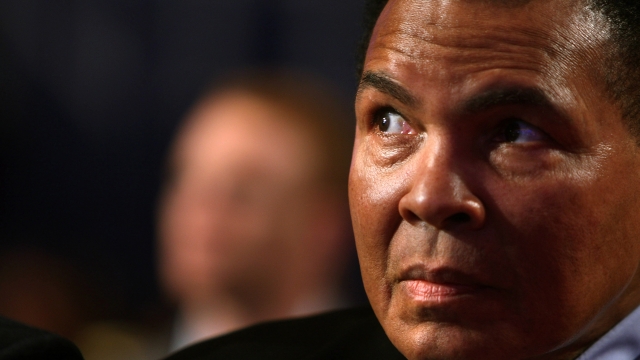 Muhammad Ali sits looking off to the side while wearing a black suit.