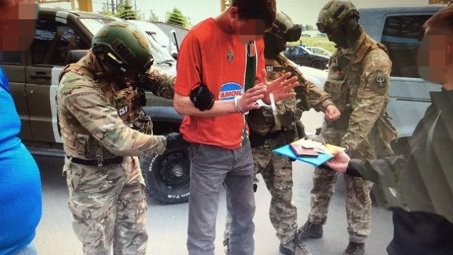 A man in an orange shirt is placed in handcuffs by security service officers wearing camouflage.