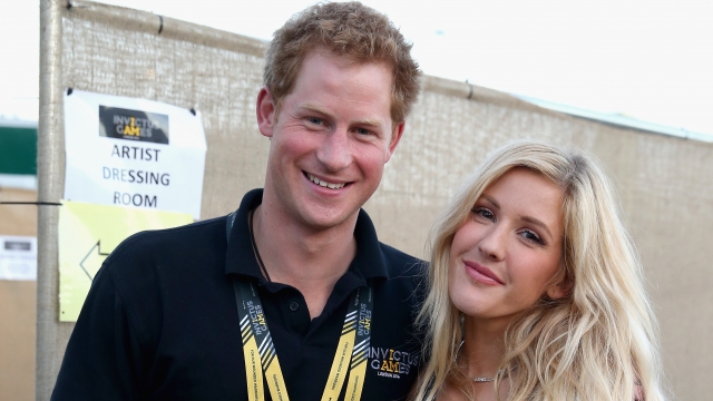 Prince Harry, wearing a black shirt, stands next to Ellie Goulding, wearing a black crop top and white pants.