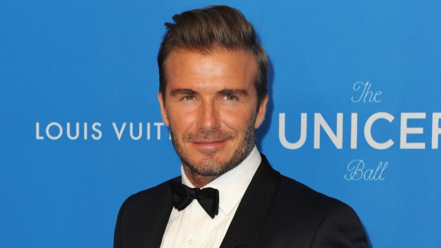 David Beckham stands in front of a blue background wearing a black tuxedo.