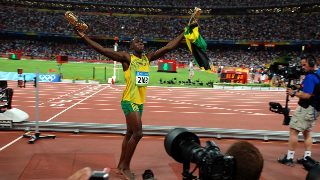 Usain Bolt in gold and green outfit holds his golden shoes and Jamaican flag in the air.