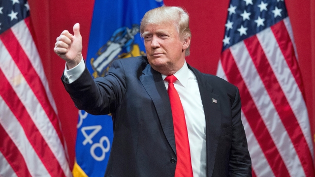 Donald Trump wearing a navy suit with white tie and red shirt give a "thumbs up" at a campaign event.