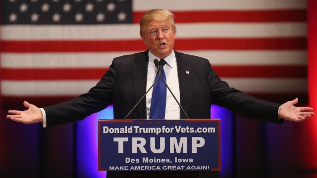 Donald Trump stands in front of an American flag backdrop with arms raised to his sides. He wears a black suit and blue tie.