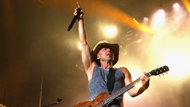 Kenny Chesney, wearing a cowboy hat, a tank top and jeans, points one hand in the air while holding a microphone.