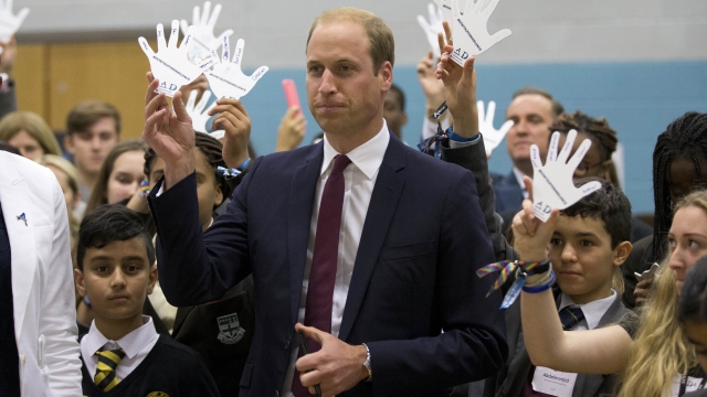 Prince William wearing a blue suit and red tie holds a paper cutout hand and is surrounded by children doing the same.