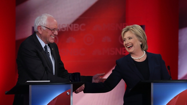 Democratic candidates Hillary Clinton and Bernie Sanders shake hands and smile at a debate.