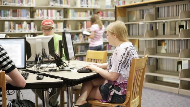 A blonde girl wearing a pink and black shirt and jean shorts sits at a black computer in a library.