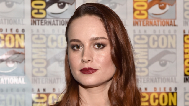 Brie Larson, with red hair, is wearing an emerald-colored jumpsuit and posing in front of a Comic-con step and repeat