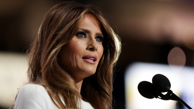 Melania Trump, wearing a white dress, stand at a podium with two microphones. She has brown wavy hair with blonde highlights.