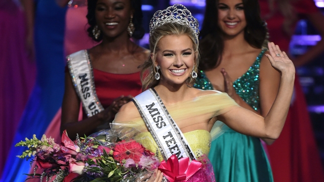 Miss Teen USA Karlie Hay, wearing a yellow floor-length gown, waves as she holds flowers and wears a crown.