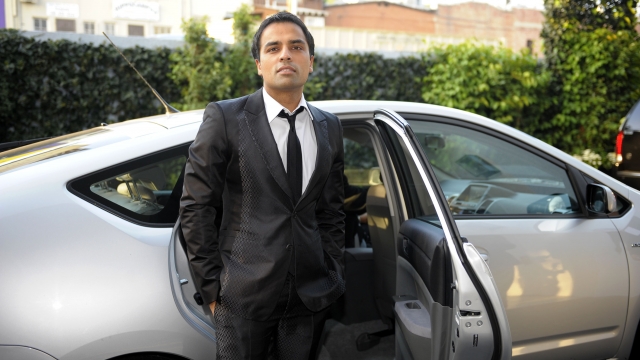 Gurbaksh Chahal stands in front of a silver sedan. He's wearing a black suit, white shirt and black tie.