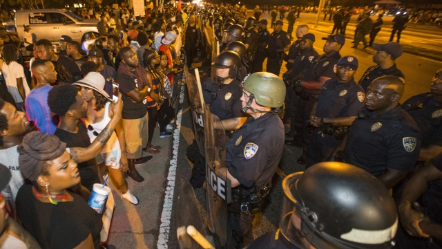 A line of protesters and police wearing riot gear meet face to face on a street.