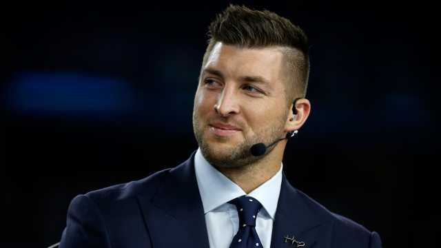 Tim Tebow, with spiked hair, wears an over the ear microphone and navy suit with navy and white tie.
