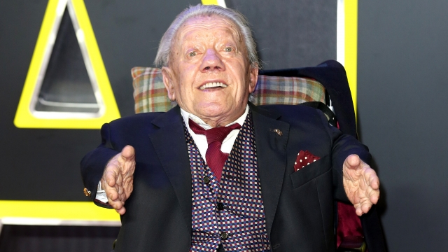 Kenny Baker sits in a wheel chair. He wears a blue suit, blue and red checkered vest and burgundy tie.