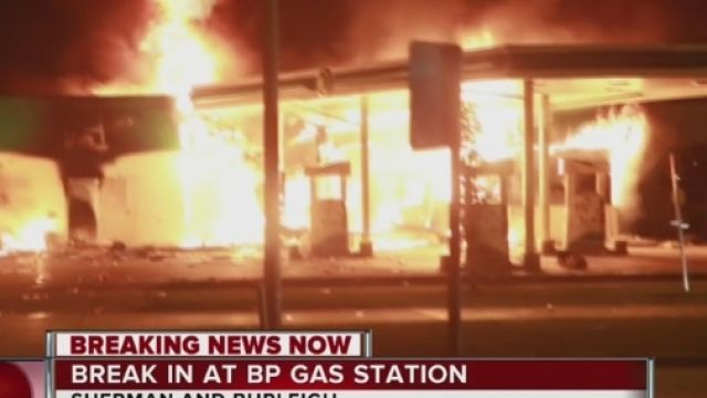 A BP gas station is engulfed in flames with flames glowing in the dark night. The building appears to be caving in.