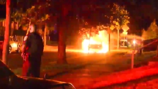 A police officer stands by his squad car as a car in the distance is engulfed in flames.