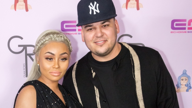 Blac Chyna and Rob Kardashian stand on a red carpet with a lavender backdrop. She wears a black gown while he wears all black