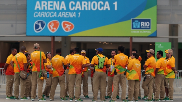 Olympic volunteers wearing orange and red shirts and khaki pants stand together outside an Olympic arena.