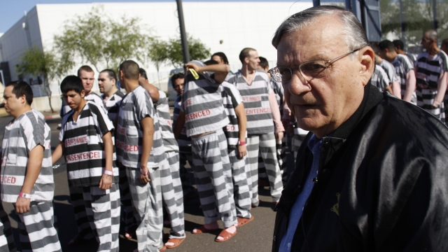 Sheriff Joe Arpaio, wears a black jacket and blue button down shirt. lnmates standing behind him wear white and black stripes