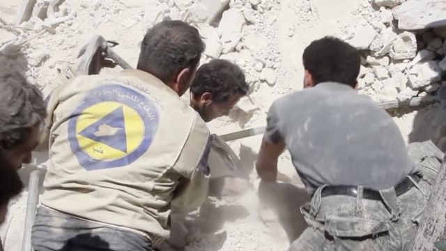 Two men wearing beige vests with a blue and yellow logo on the back appear to dig through rubble.