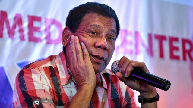 Philippine President Rodrigo Duterte holds his hand to his cheek and holds a microphone. He is wearing a red, plaid shirt.