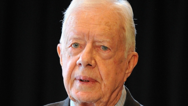 Former President Jimmy Carter stands at a podium with a microphone. He has white hair and wears a gray suit with red tie.