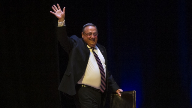 Gov. Paul LePage, wearing a suit, walks on stage waving his right hand in the air. He carries a portfolio in his left hand.