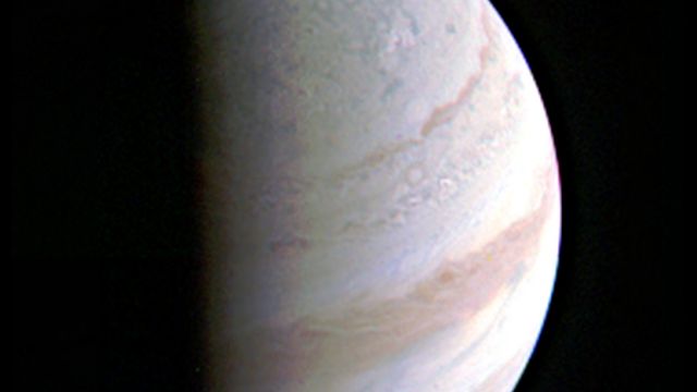 Jupiter, the big planet, is covered in a dark shadow on its left side, and has streaks of orange and white.