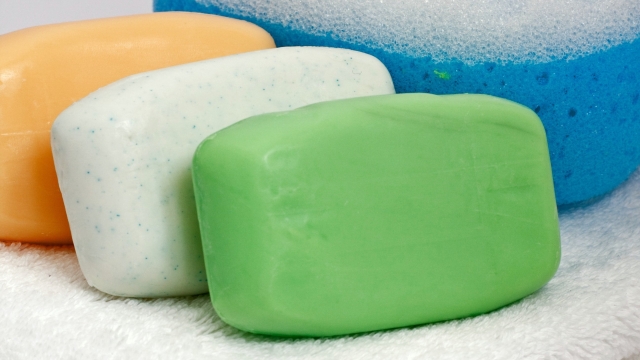 Three bars of soap, one white, one green, one orange sit on top of a white towel next to a blue scrubber with white bristles.