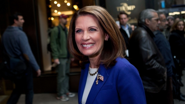 Michele Bachman smiles while onlookers stand behind her. She wears a blue jacket, with a white shirt and American flag pin.