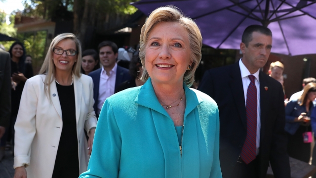 Hillary Clinton wears a blue suit. She walks by the camera and smiles followed by a member of the secret service.
