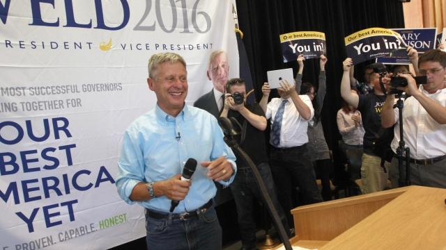Gary Johnson wears a blue and white checkered button-down shirt. He holds a black microphone and smiles.