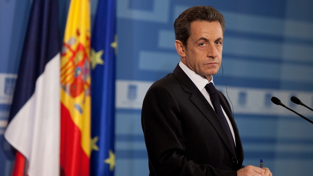Former French President Nicholas Sarkozy stands at a podium looking to his right. He wears a black suit and black tie.