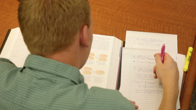 An overheard view of a man wearing a green shirt sits at a table with a textbook and notebook in front of him.