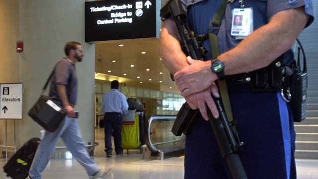 A shot of an airport security officer from the waist down holding a gun. A man rolling a suitcase walks in the background.