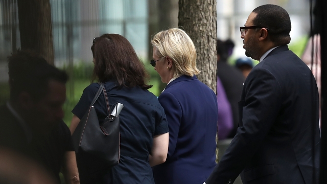 Hillary Clinton wearing a navy suit and dark sunglasses as she walks away and close to a woman in a black short-sleeved shirt