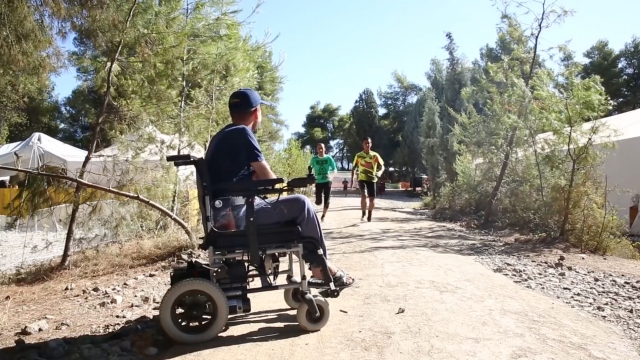A Syrian refugee with muscular dystrophy sits in a wheelchair as two people run toward him.