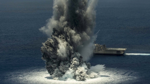 A massive explosion from the ocean with a navy ship behind it. The explosion is white towards the surface and dark gray above
