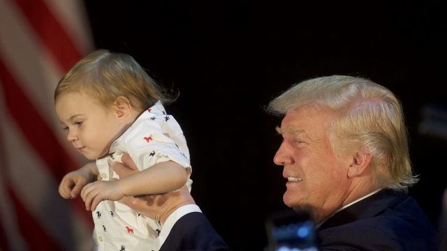 Donald Trump, wearing a navy suit and white shirt, is viewed from the side. He holds a baby boy wearing a white collar shirt.