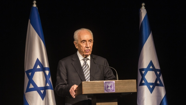 Shimon Peres stands at a podium with a microphone between two Israeli flags.