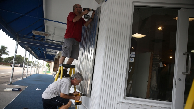 A man stands on a ladder drilling storm shutters onto windows. Another man kneels while boarding up the windows.