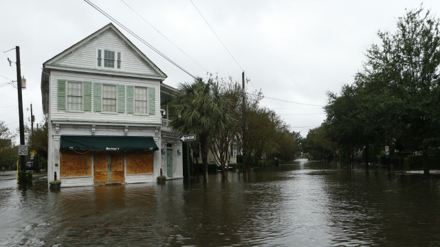A house with boards in the bottom-floor windows sits on a flooded street.
